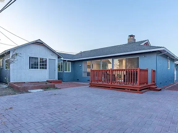 Blue house with a wooden front deck and large paver driveway at sunset