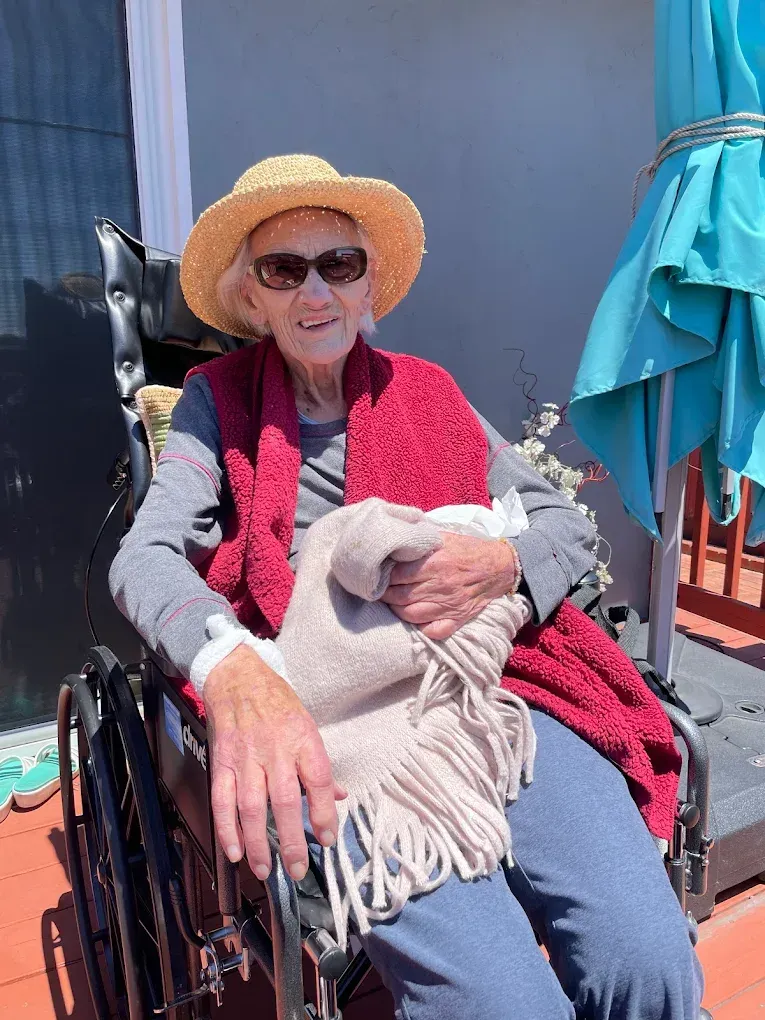 Person in a wheelchair wearing a straw hat, red vest, and sunglasses, holding a light blanket outdoors.
