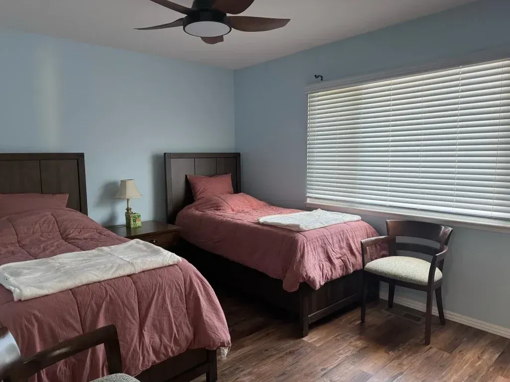 Two twin beds with pink bedding in a light blue bedroom with a ceiling fan and chair by the window.