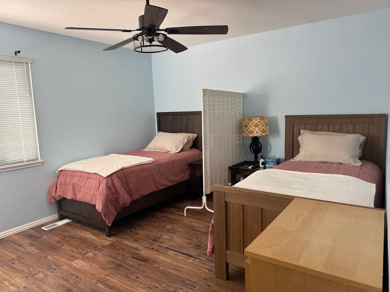 Two twin beds in a light blue bedroom with a ceiling fan, nightstand, lamp, and window blinds.
