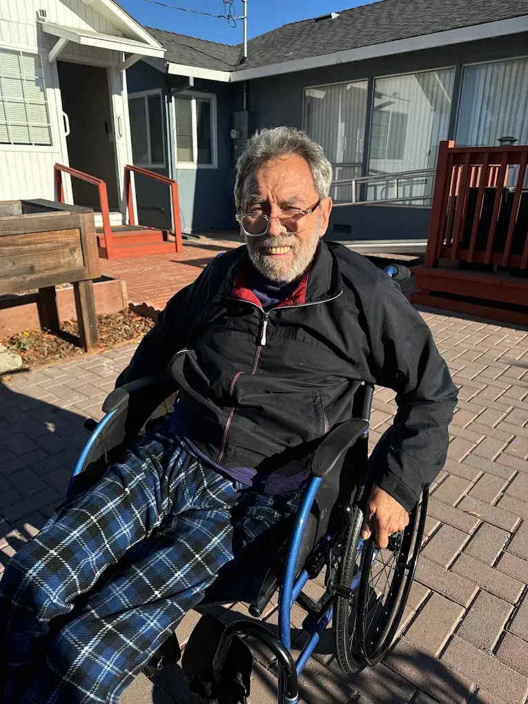 Man in wheelchair sitting outdoors in a sunny courtyard, wearing a black jacket and blue plaid pants.
