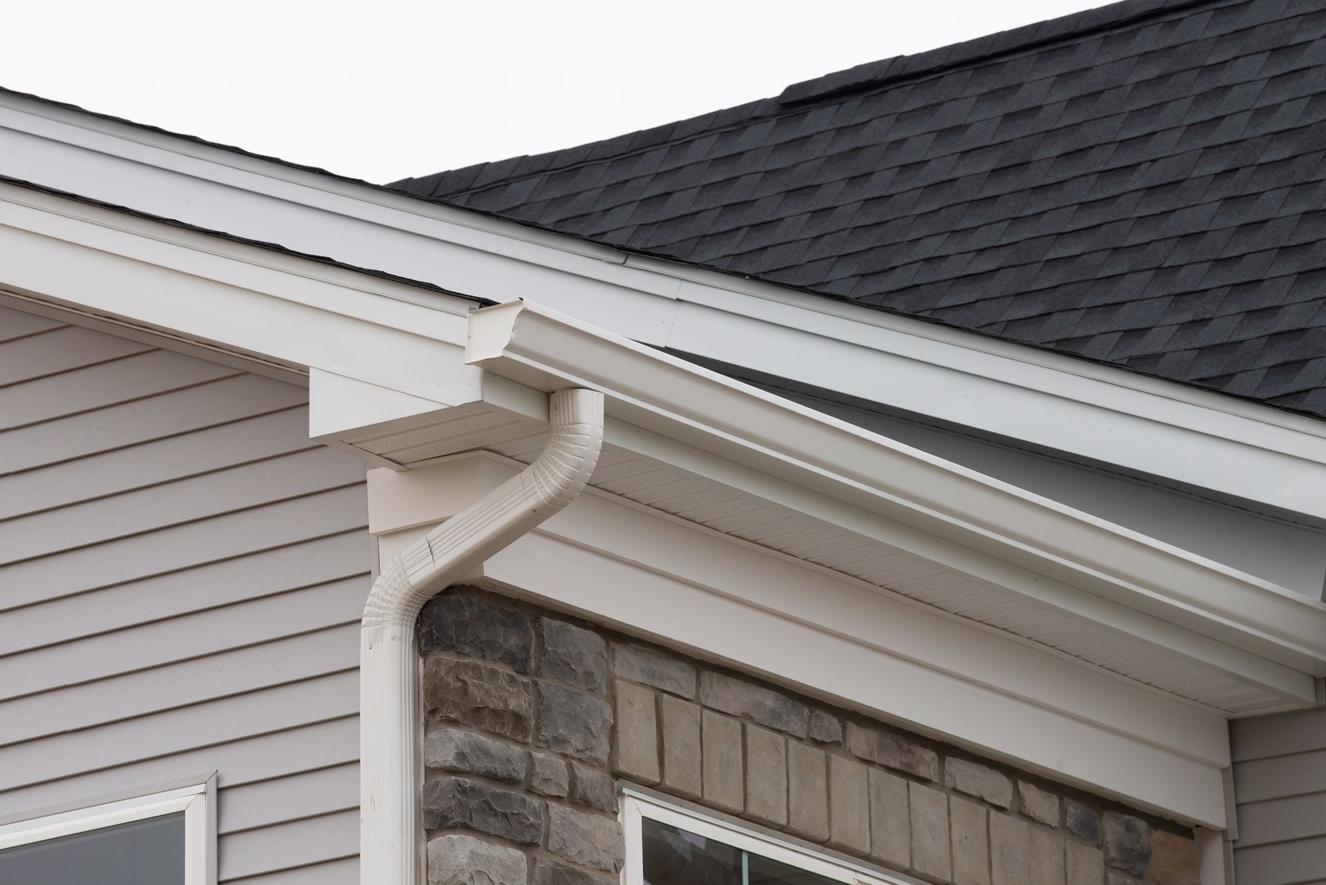 Gouttière et descente pluviale blanches fixées à l'angle d'une maison avec un revêtement gris clair, un parement en pierre