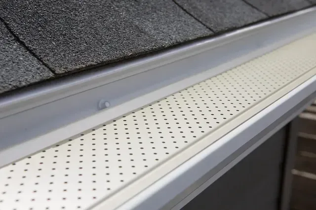 Une grille de protection de gouttière en métal perforé de couleur claire, installée sur une gouttière de toit