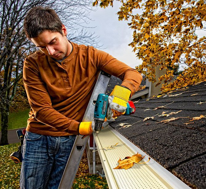 Un homme sur une échelle nettoie une gouttière avec un outil électrique ; on aperçoit des feuilles d'automne.
