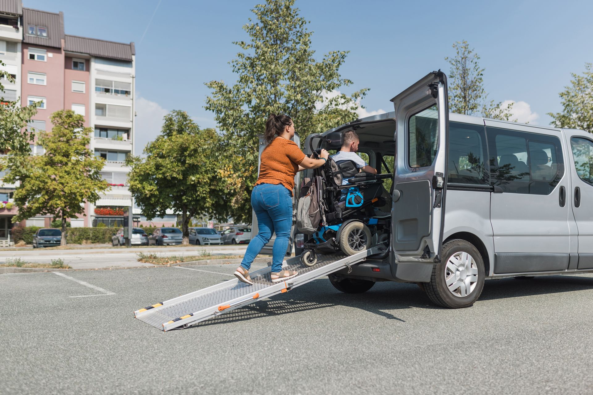 Two people loading a wheelchair into a van using a ramp in a parking lot