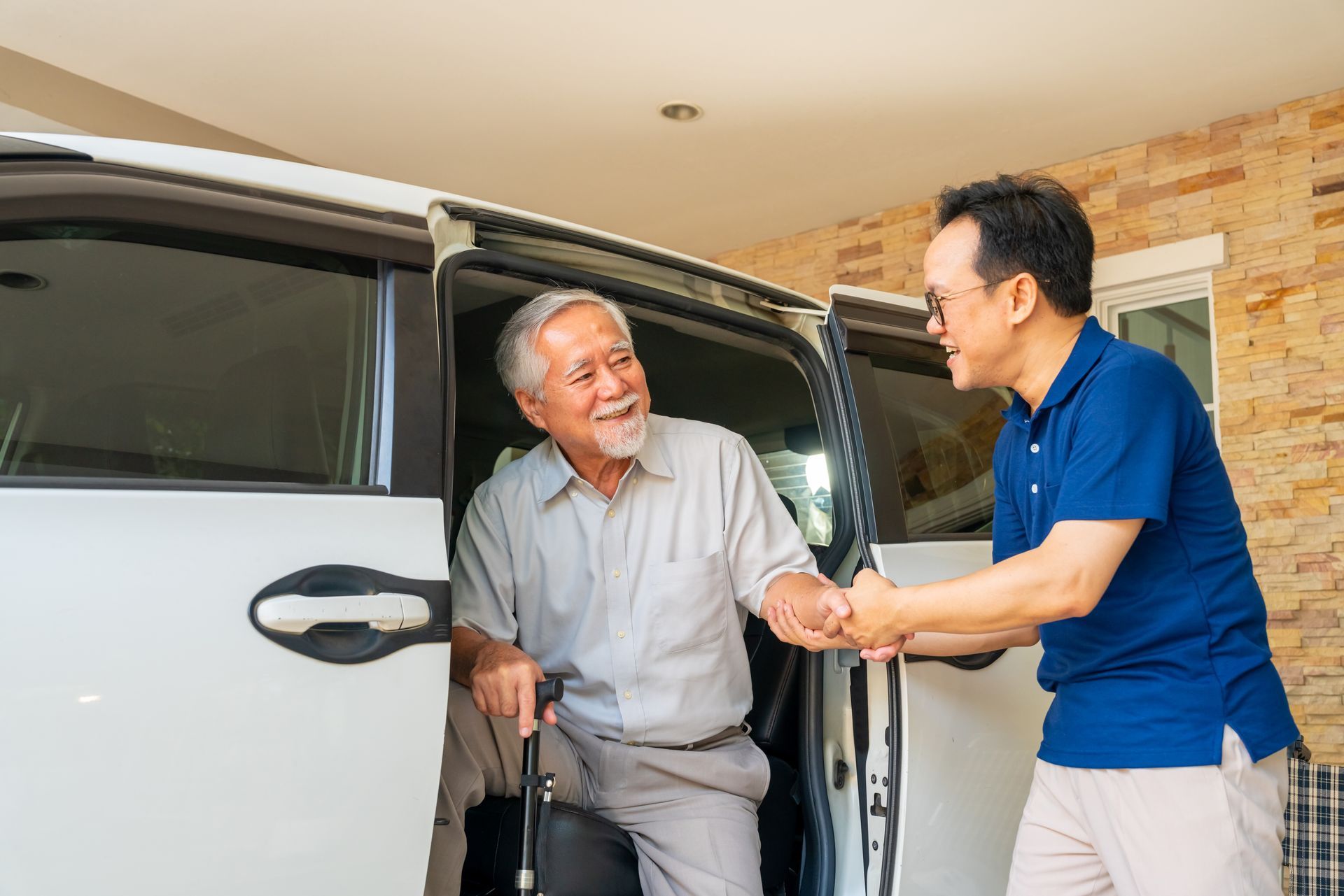 Man helping an older man out of a car in a driveway, both smiling