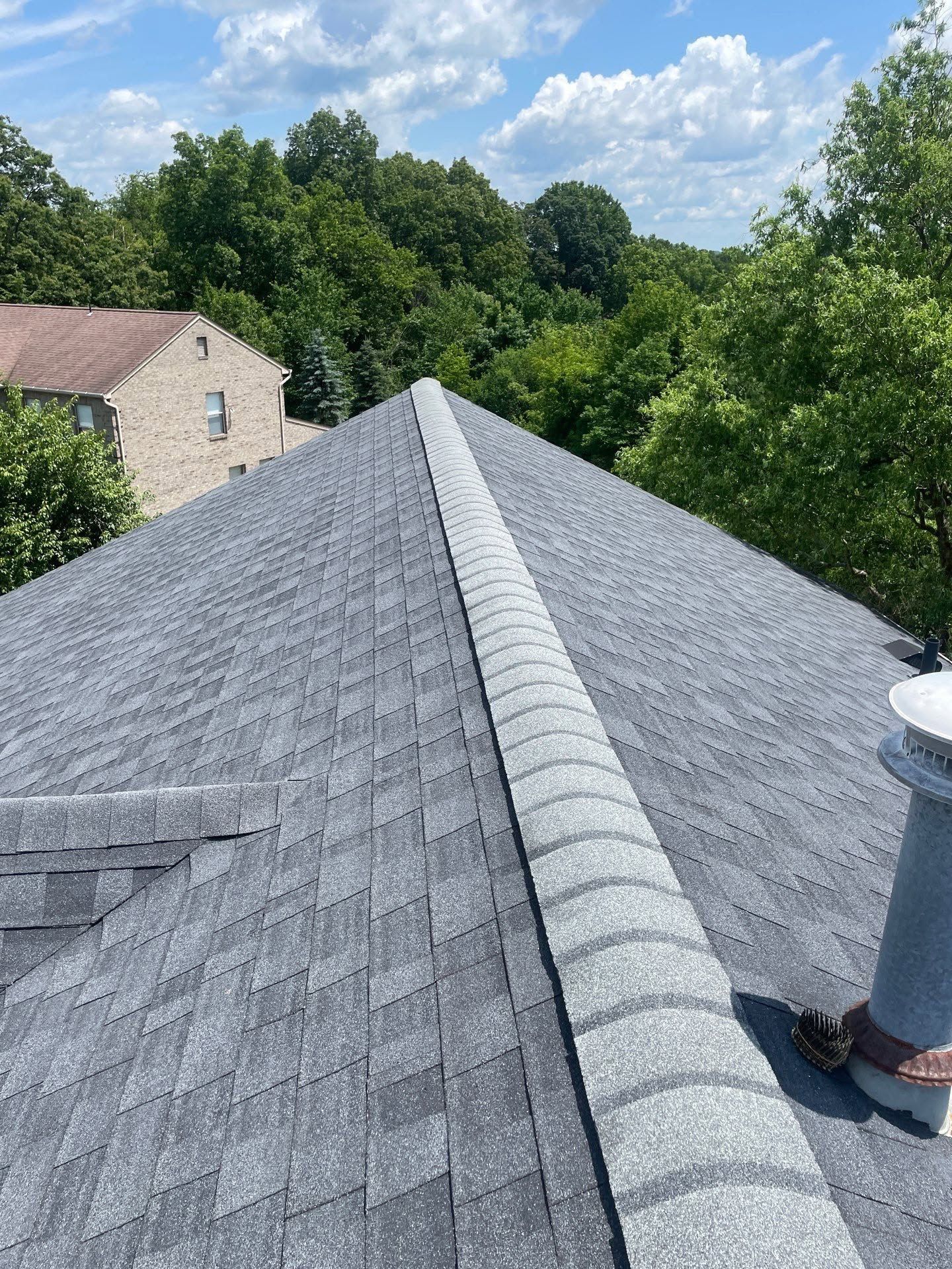 Roof ridge with gray shingles beside green trees under a blue sky, viewed from the rooftop