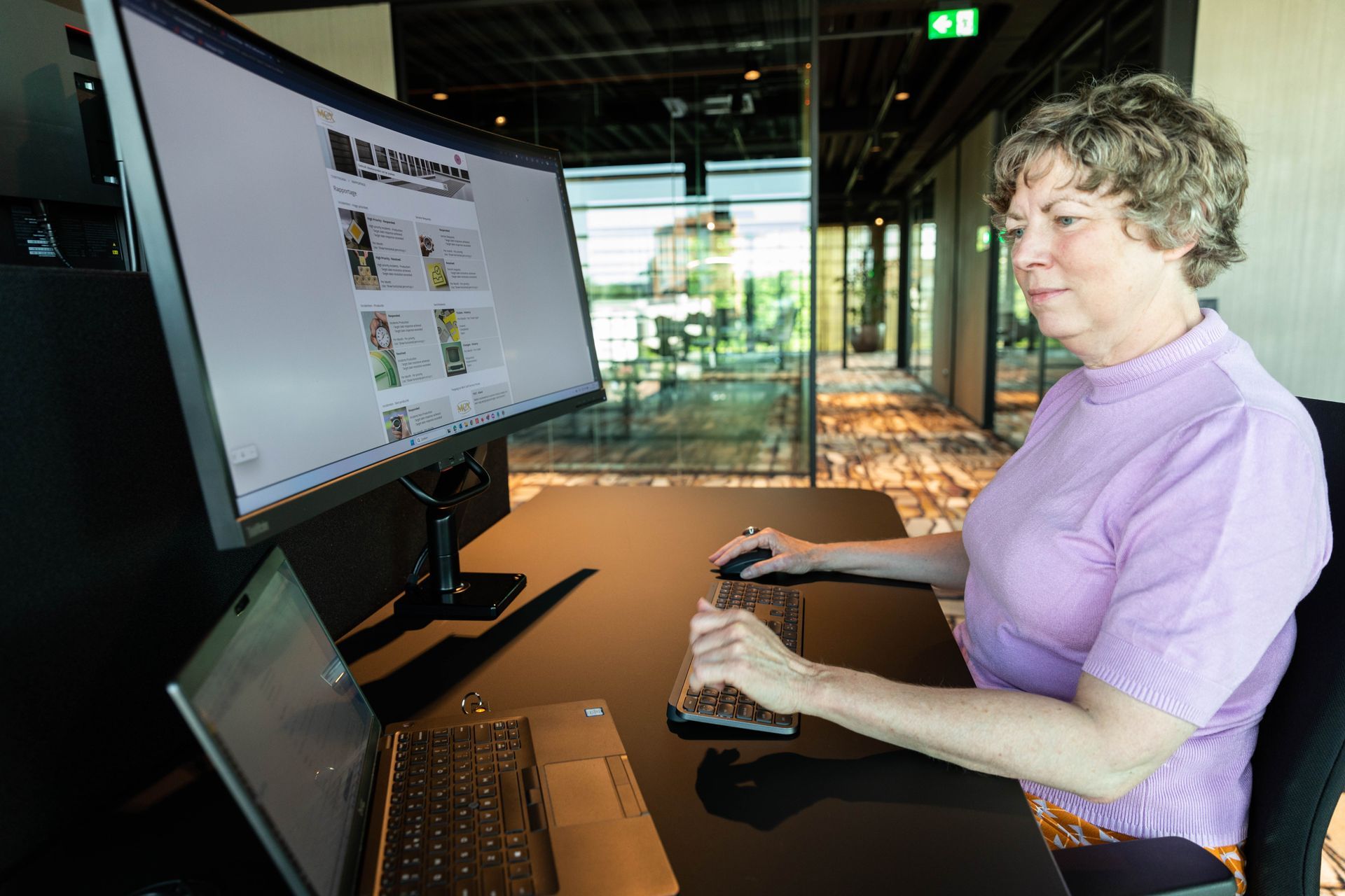 Vrouw aan een bureau, gebruikmakend van een toetsenbord en muis naast een grote monitor in een modern kantoor.