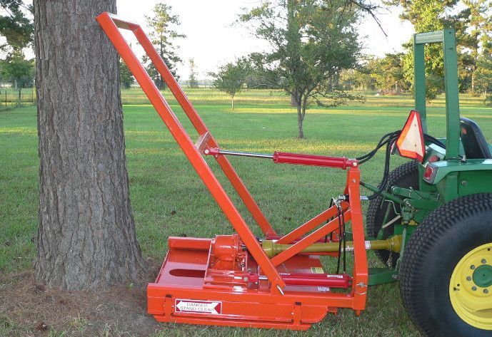 Orange tree shear attached to a green tractor, cutting a tree in a grassy field.