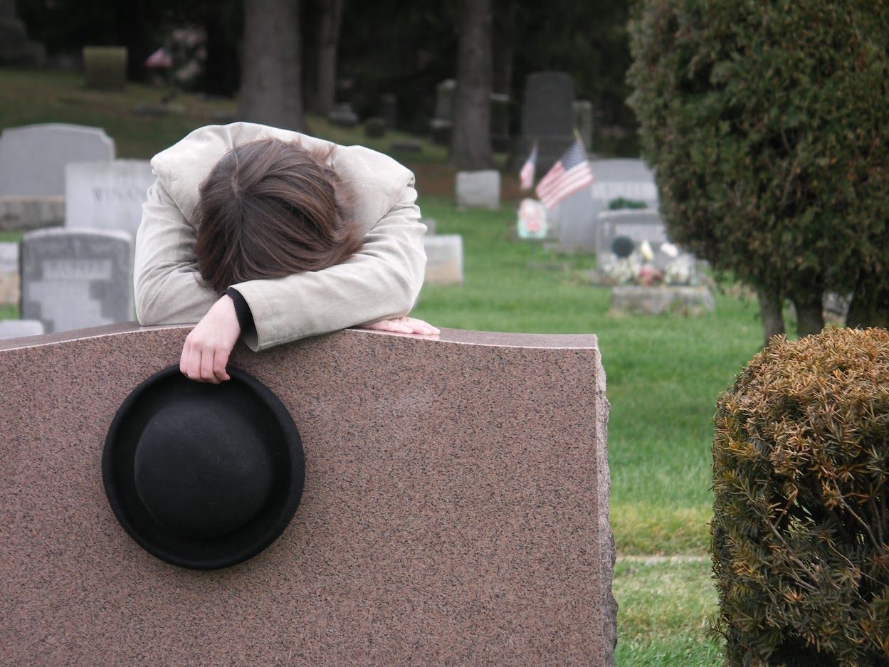 Person in a beige jacket weeping at a grave, holding a hat. Cemetery setting, overcast.