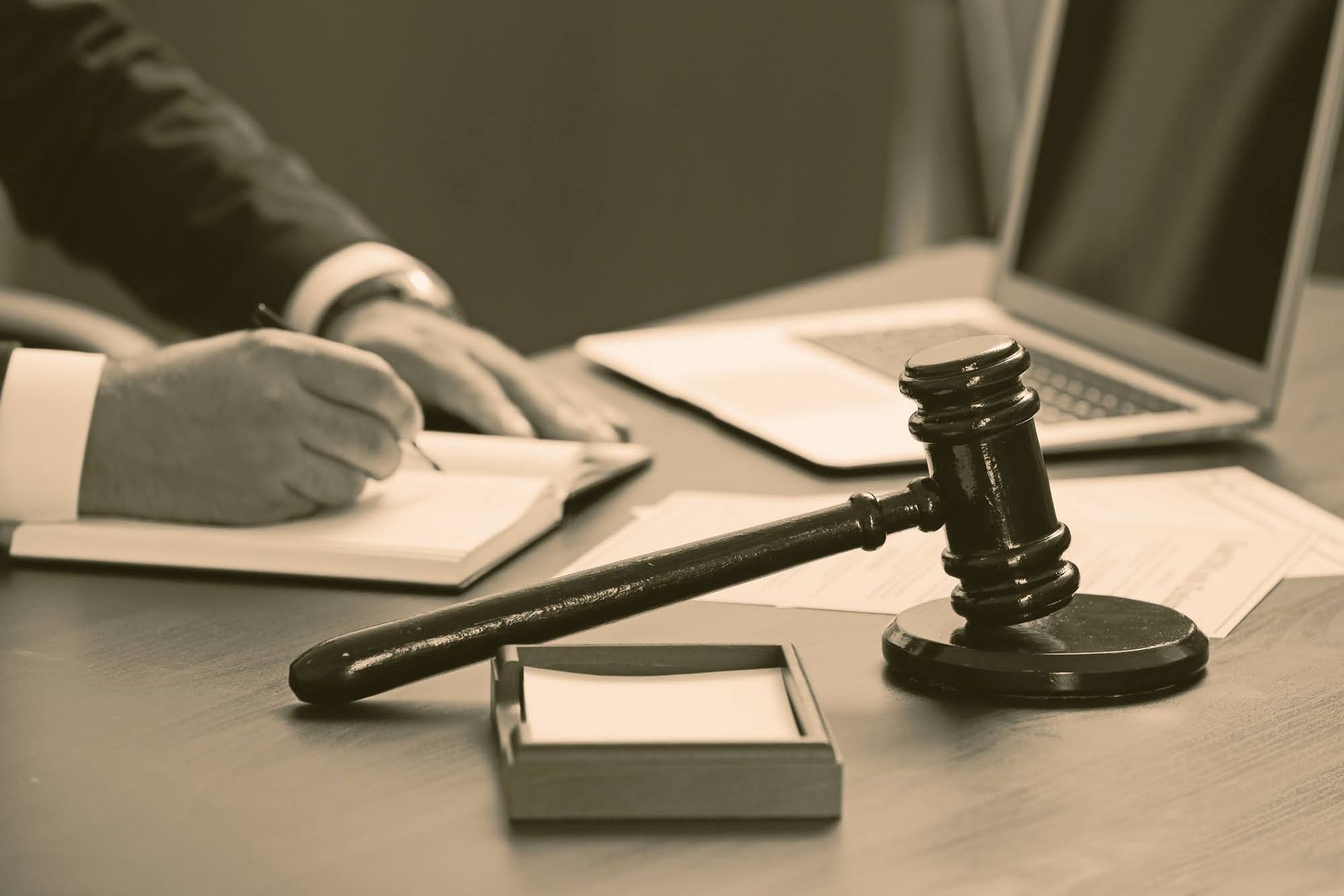A gavel rests on a desk next to a person writing in a notebook, with a laptop in the background.