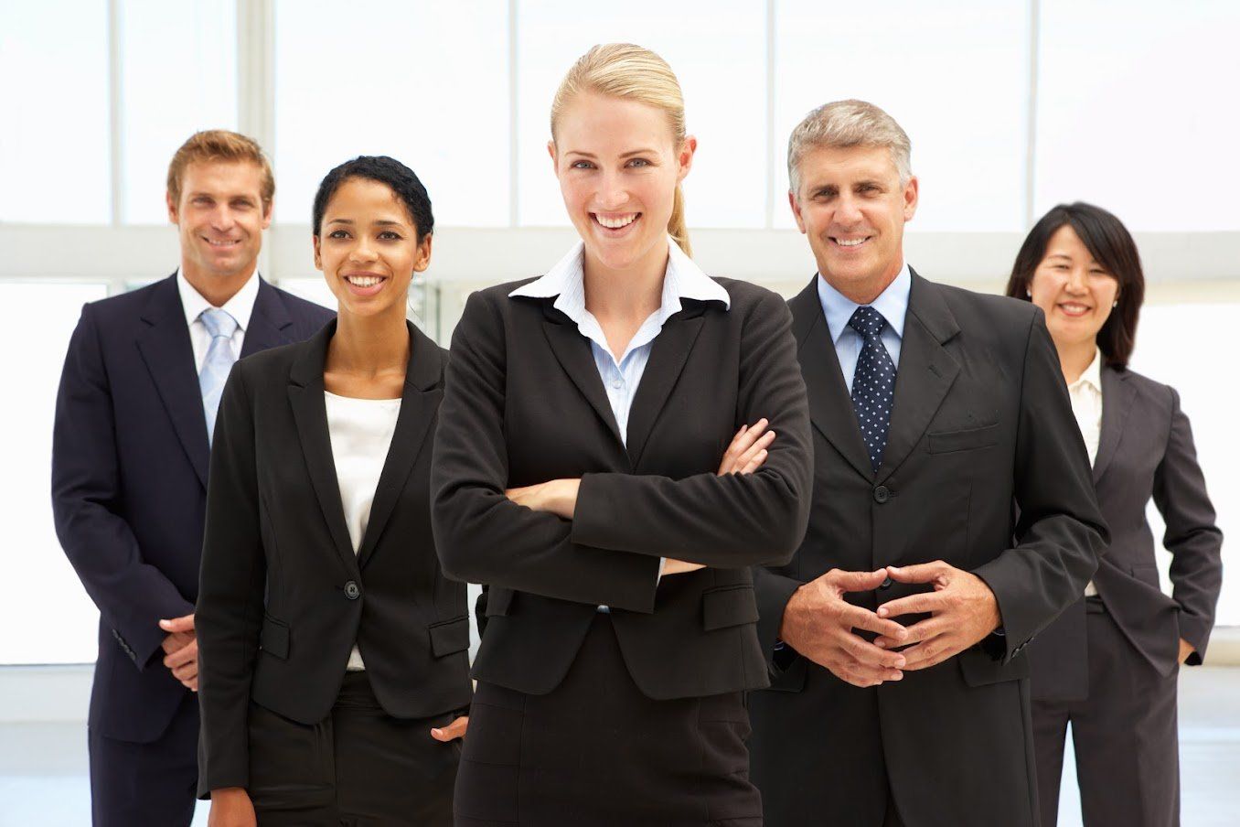 Group of five diverse business professionals in suits, smiling in an office setting.