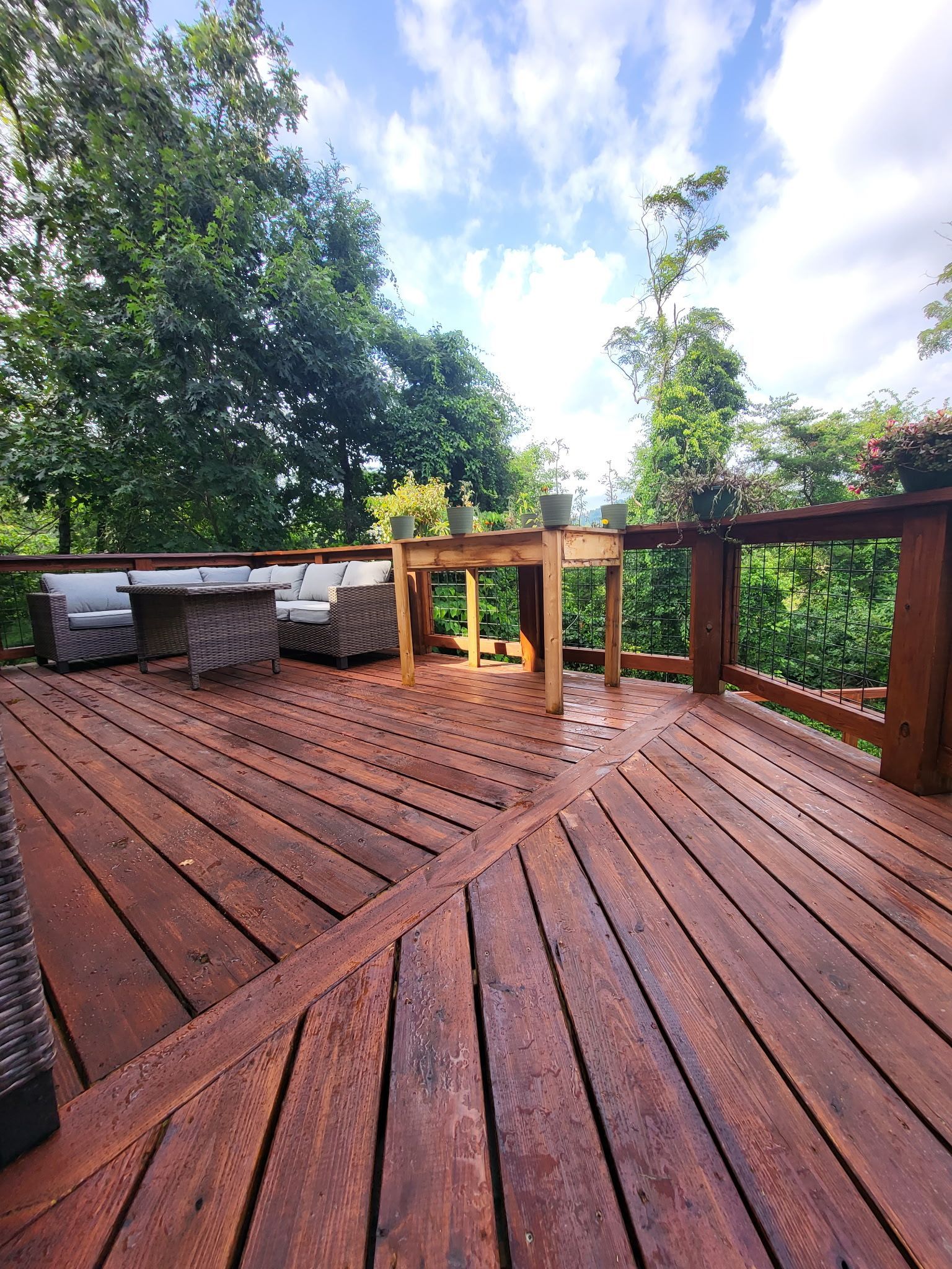 Wooden deck with railing, outdoor seating, and trees under a partly cloudy sky