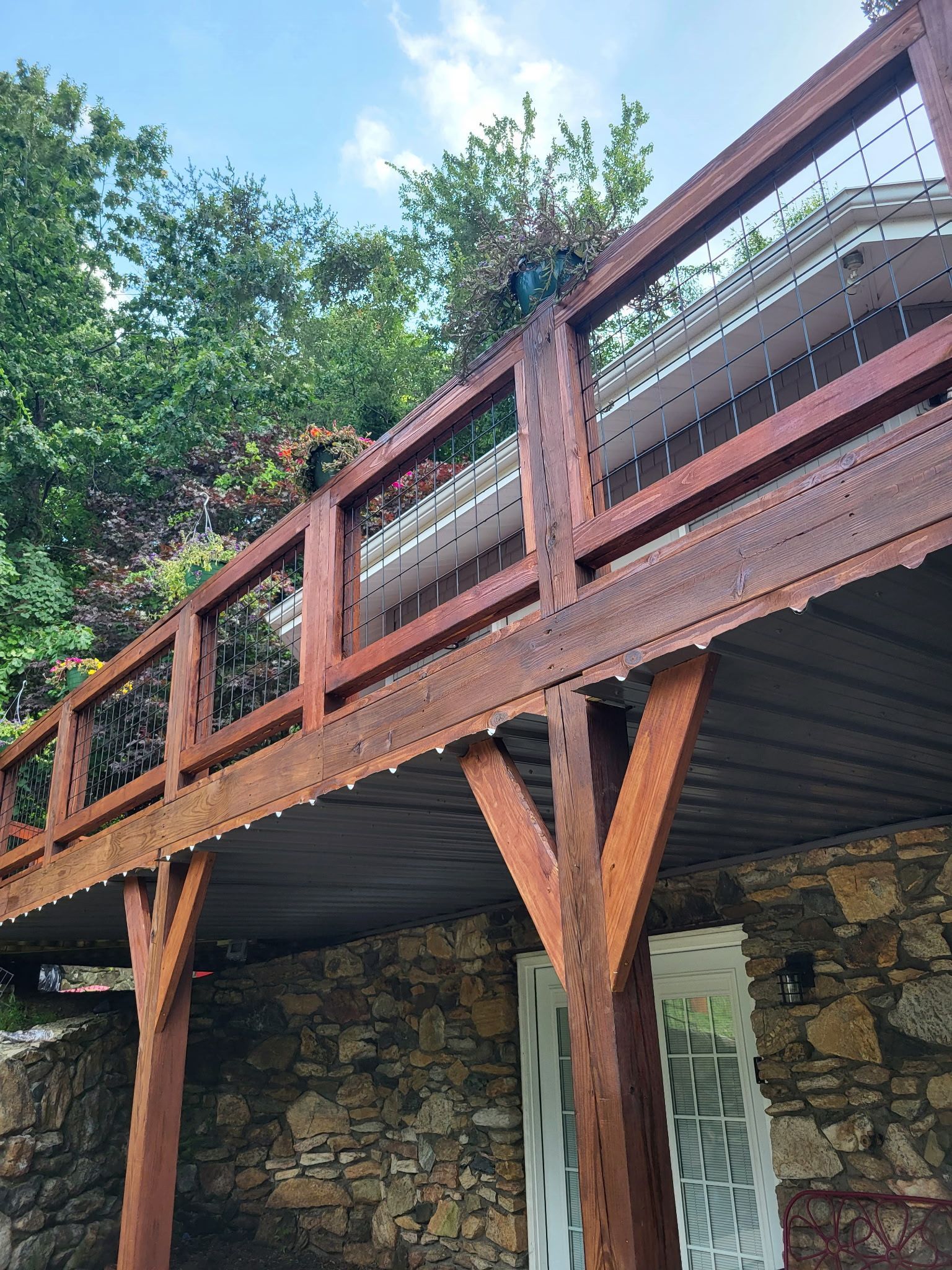 Wooden deck with railing and support posts above a stone building, framed by leafy trees.