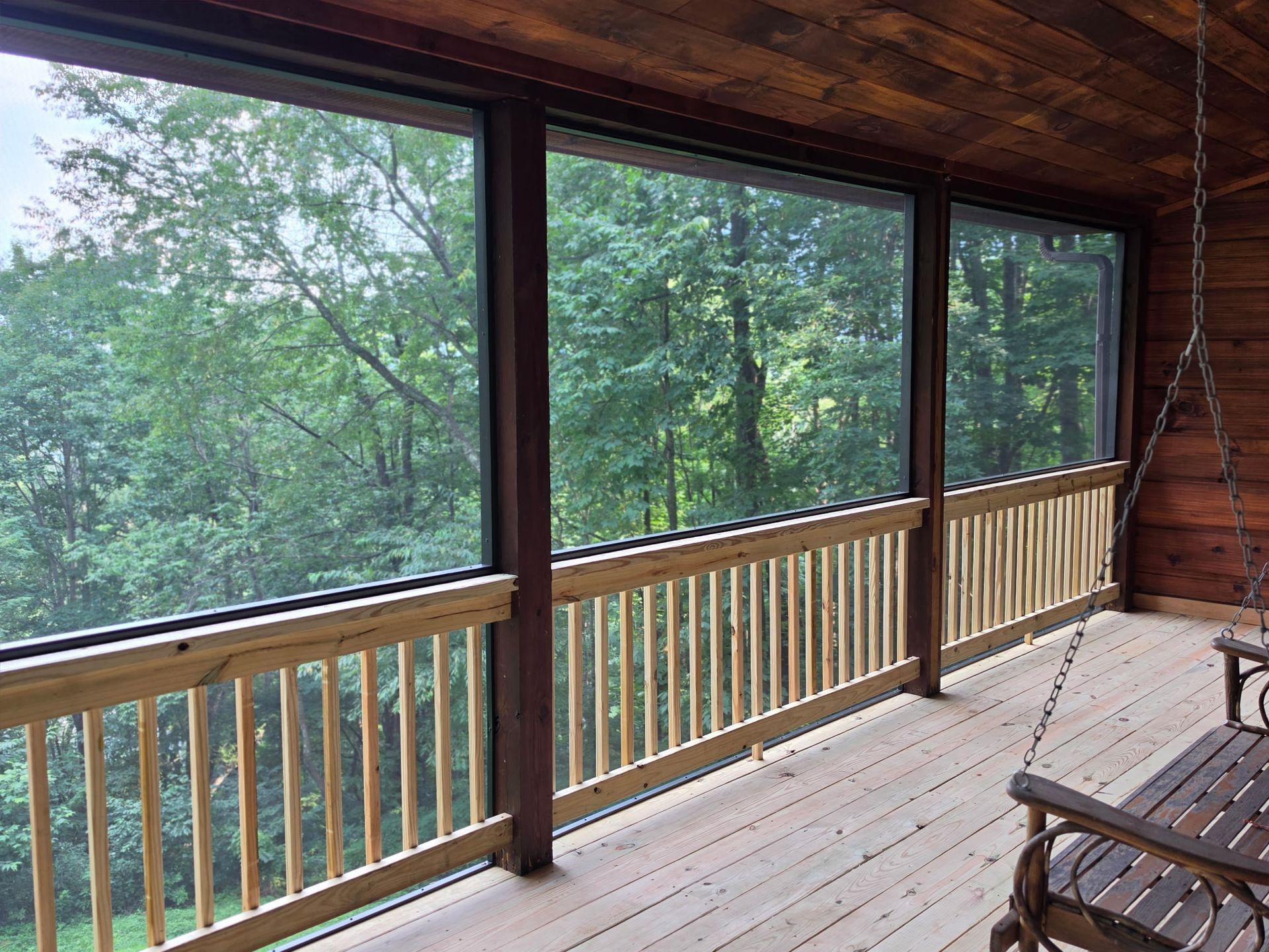 Covered wooden porch with screened windows overlooking a leafy forest; chairs sit on the deck.