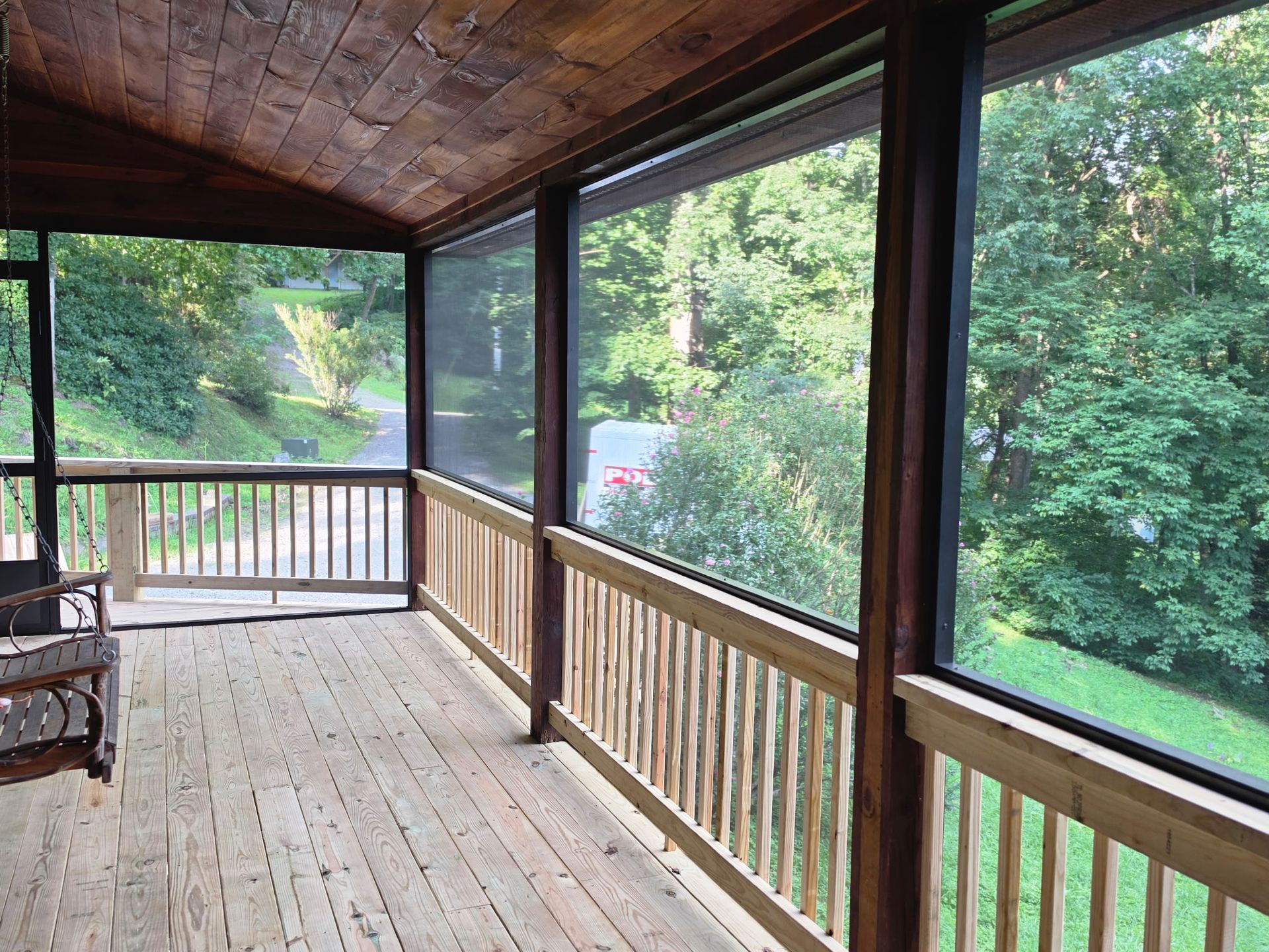 Covered wooden porch with screened windows overlooking green trees and a deck railing
