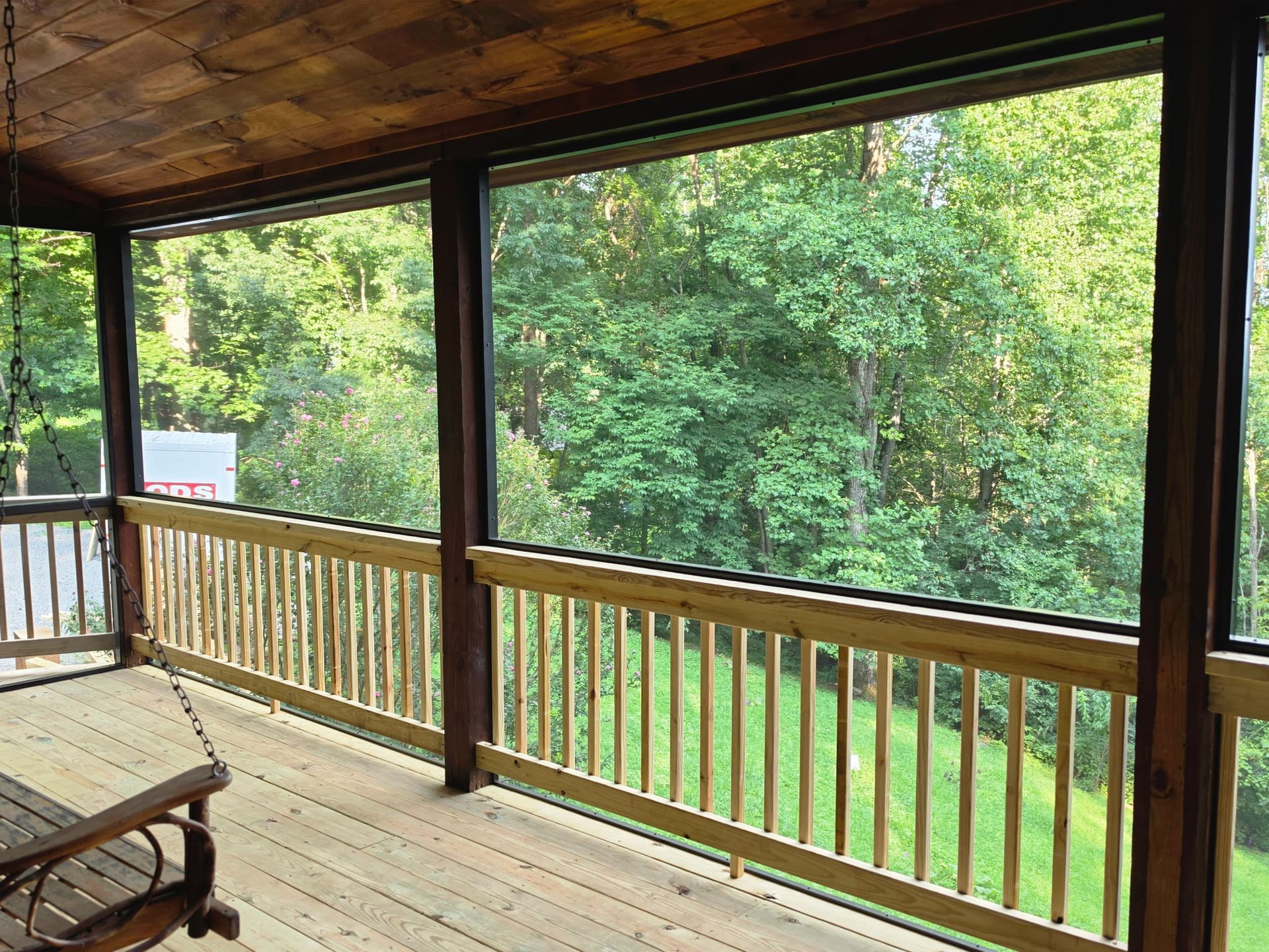 Covered wooden porch overlooking green yard and trees, with railing and bench swing.