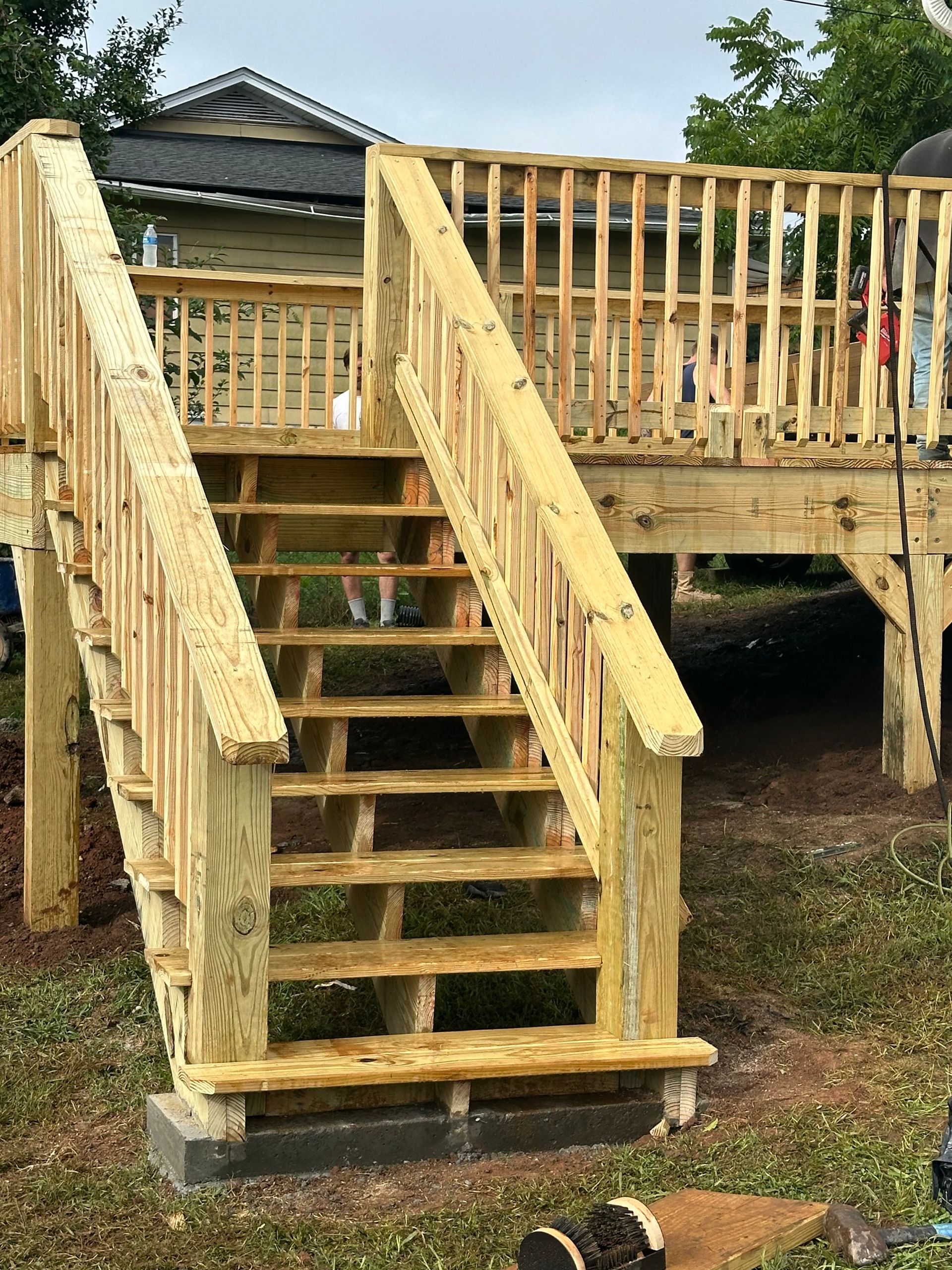 Wooden outdoor stairs and deck under construction on concrete blocks, with railings and a house in the background