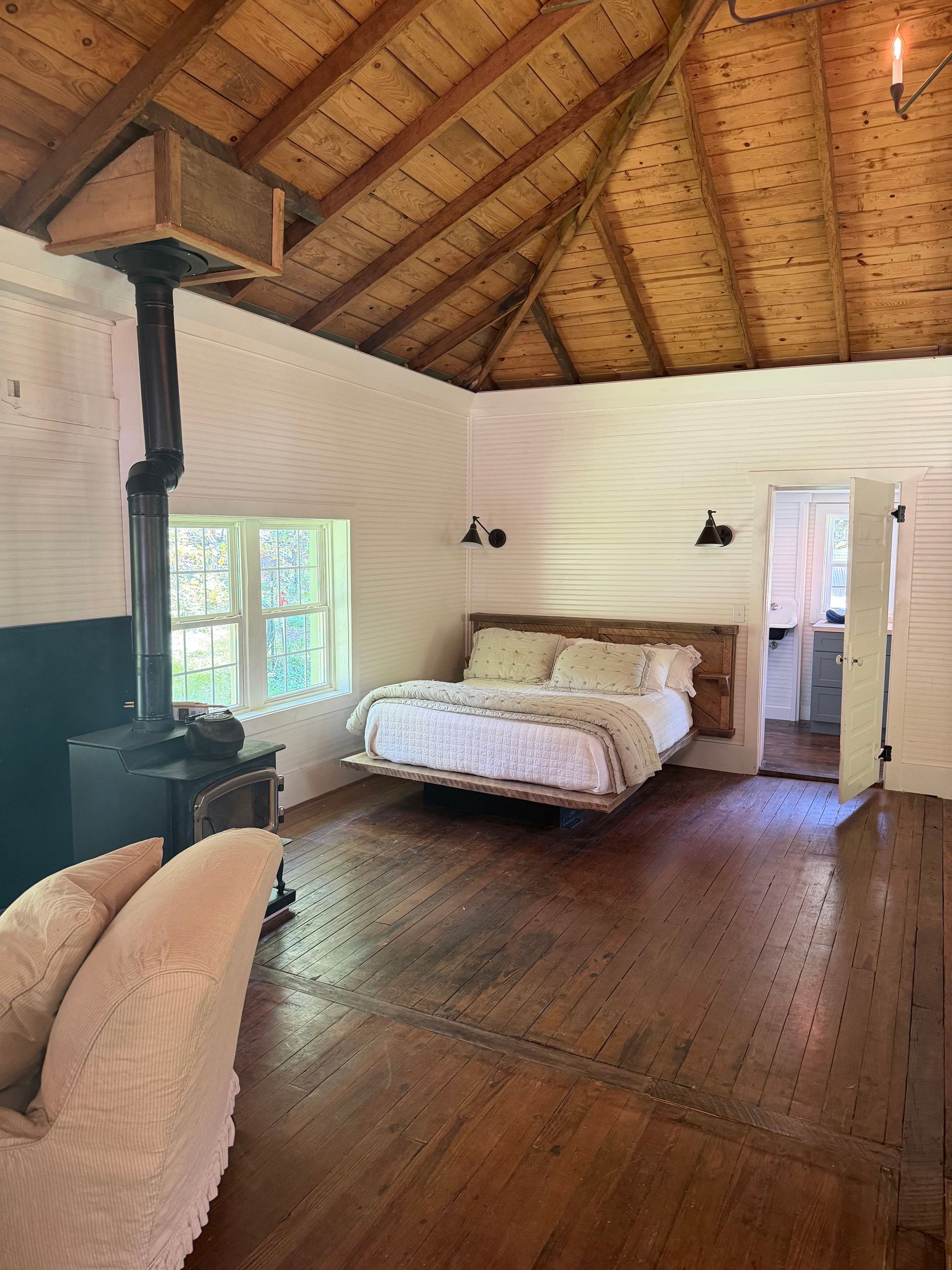 Rustic bedroom with vaulted wooden ceiling, bed, wood stove, and open doorway