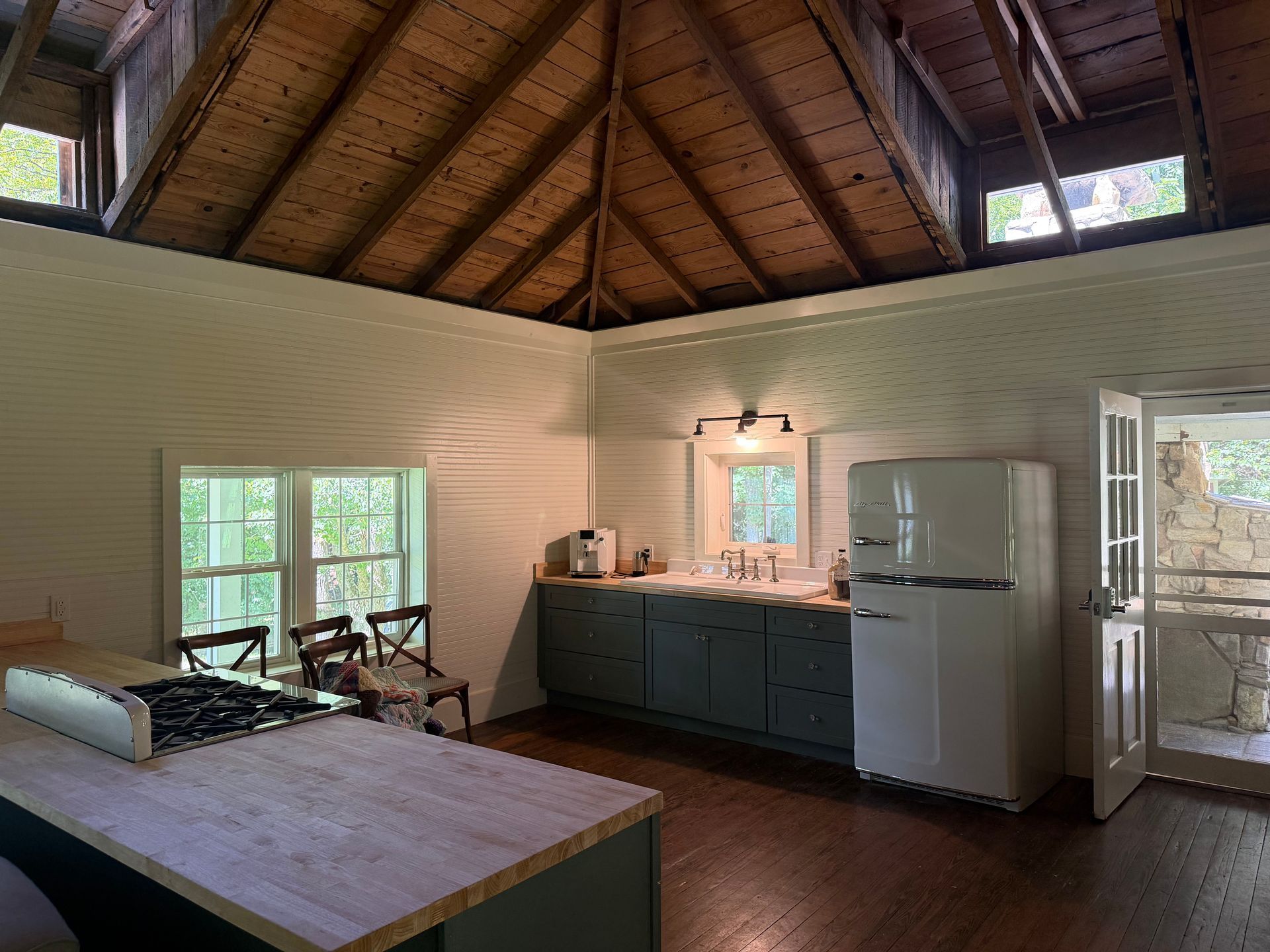 Small cabin kitchen with wood ceiling, white fridge, blue cabinets, and large windows.