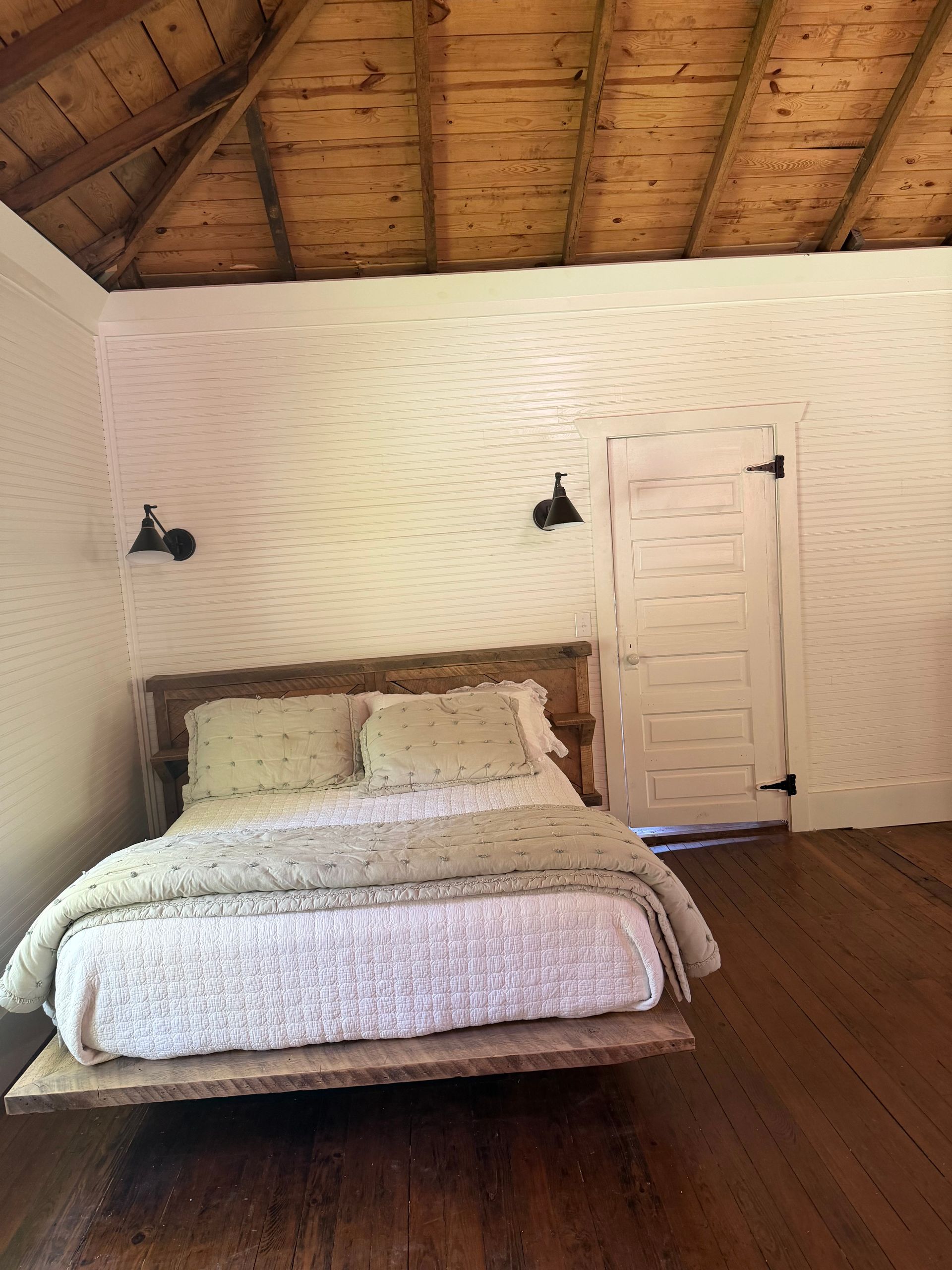 Cozy bedroom with a wooden vaulted ceiling, white walls, a bed, two wall lamps, and a closed door