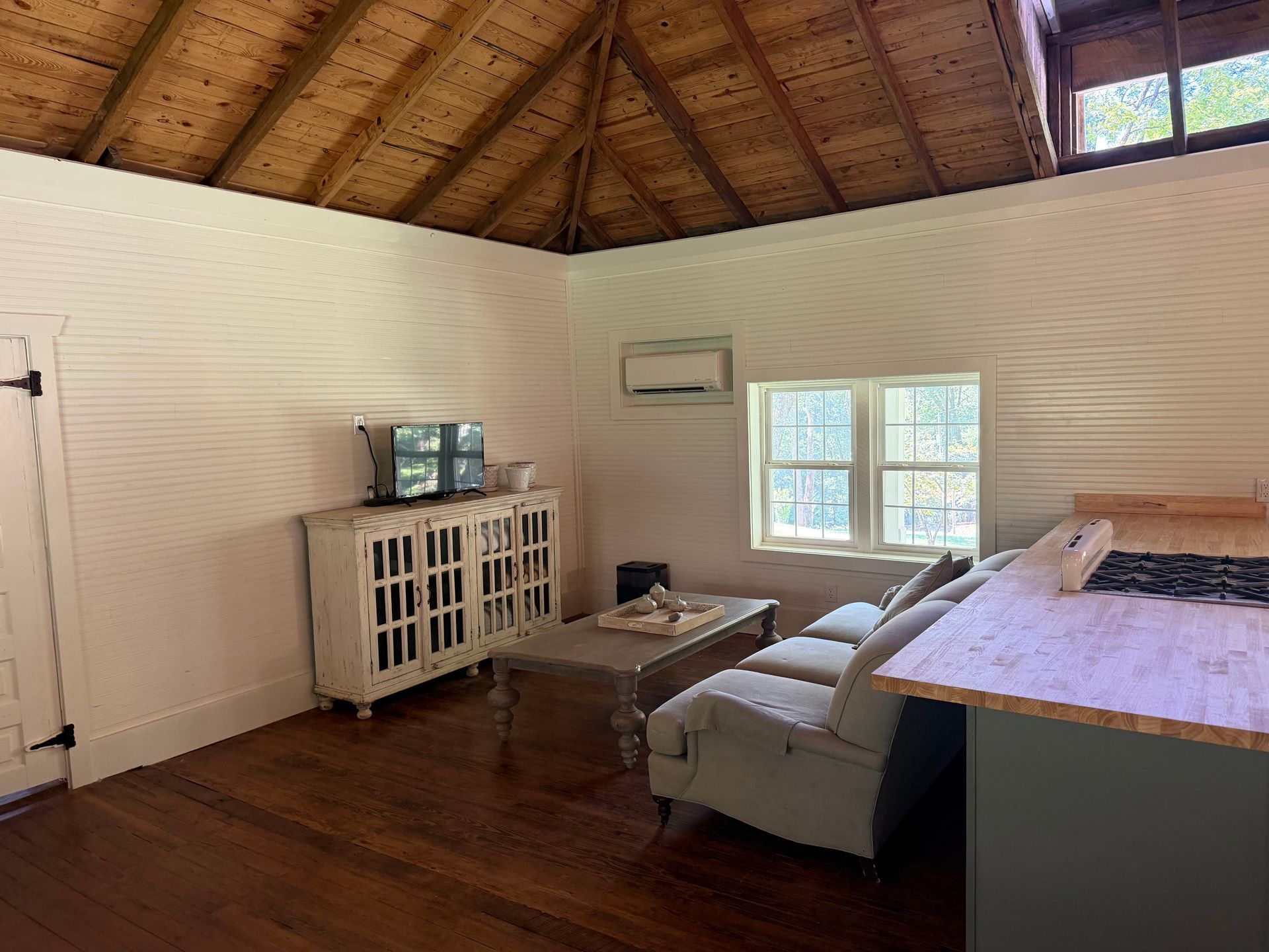 Cozy loft living area with wood-paneled walls, gray sofa, TV cabinet, windows, and vaulted wooden ceiling