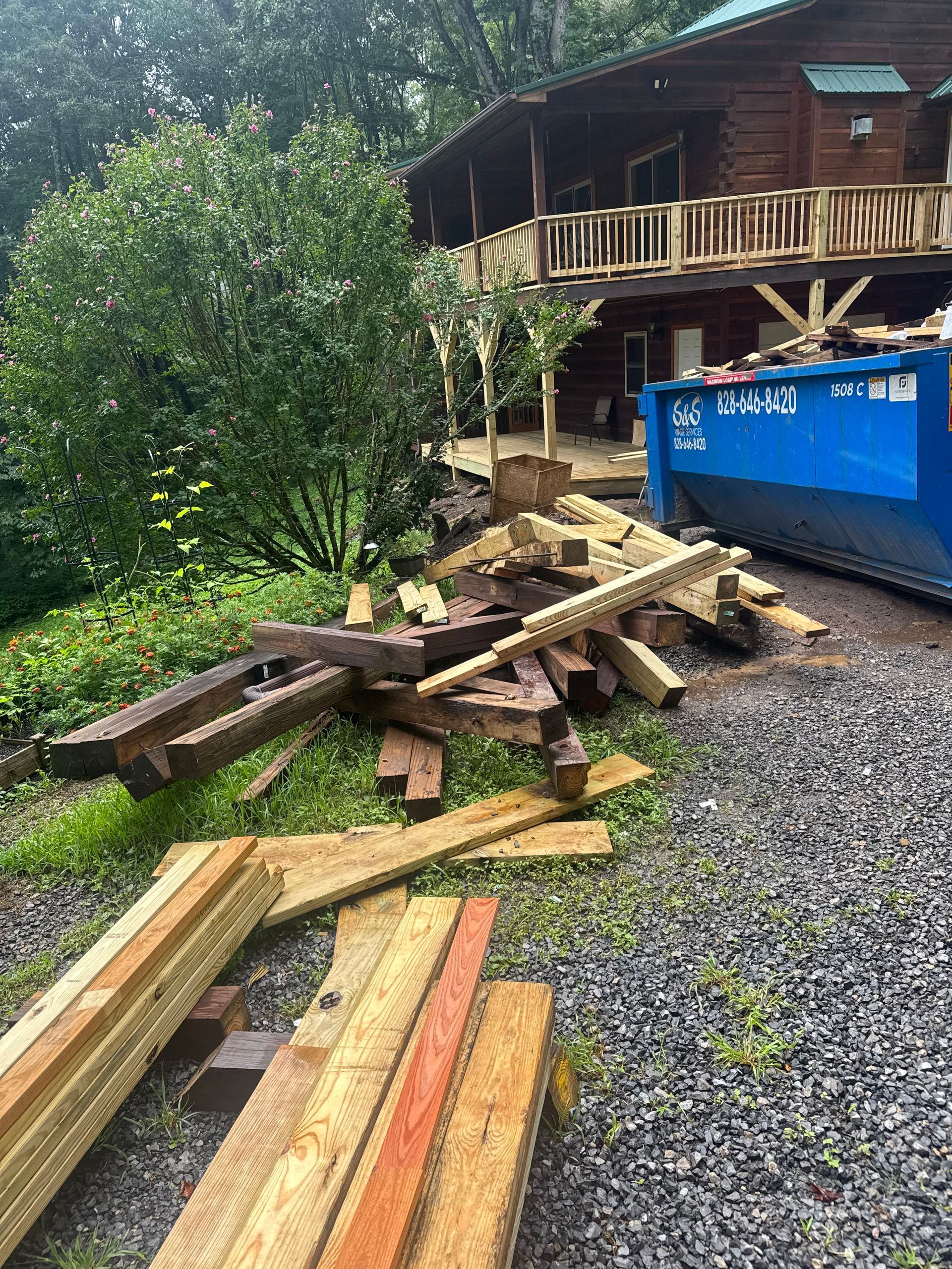 Pile of lumber and wooden beams beside a blue dumpster near a house under construction