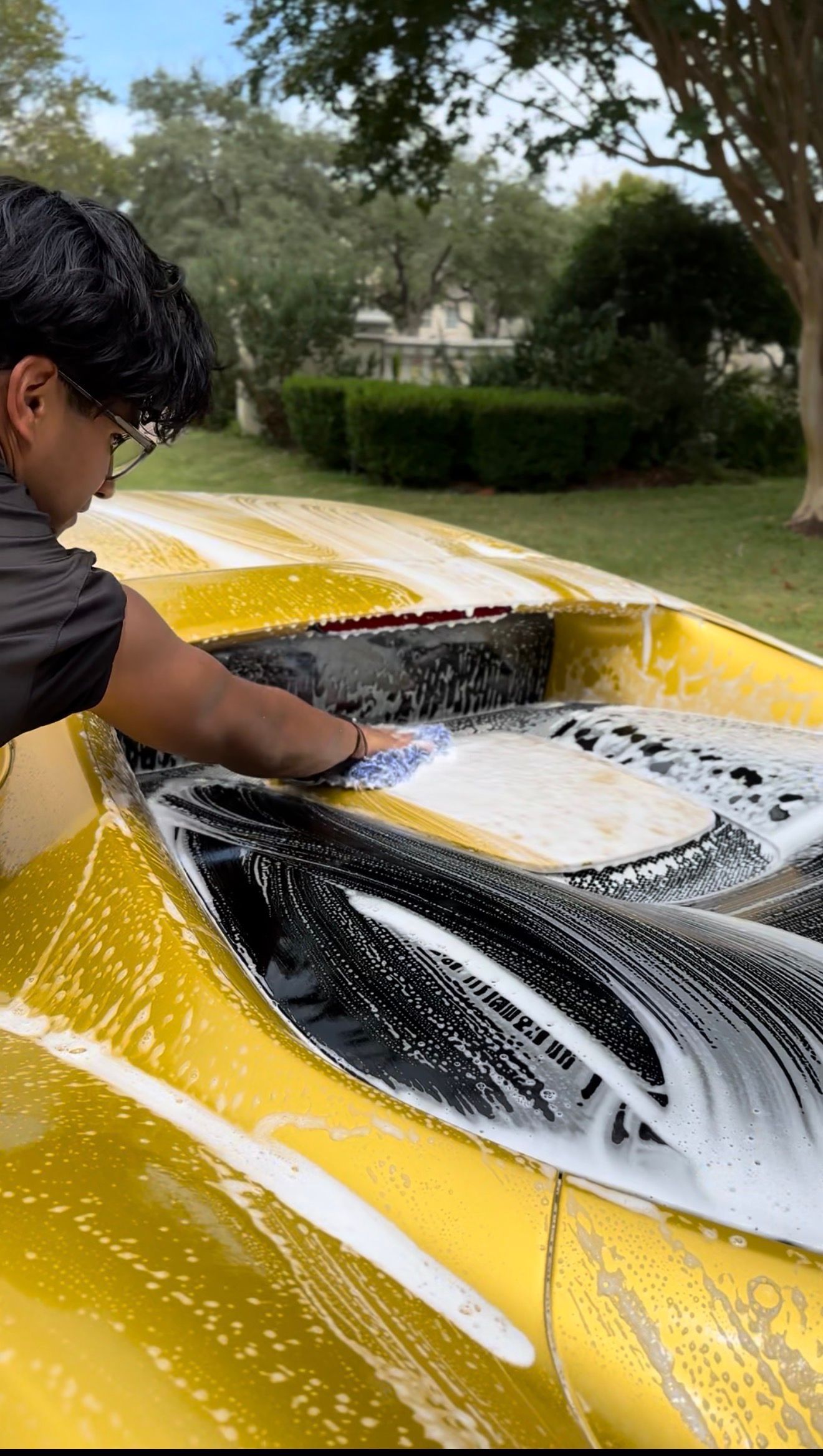A person washes the rear engine cover of a yellow sports car with a soapy sponge in a grassy outdoor setting.