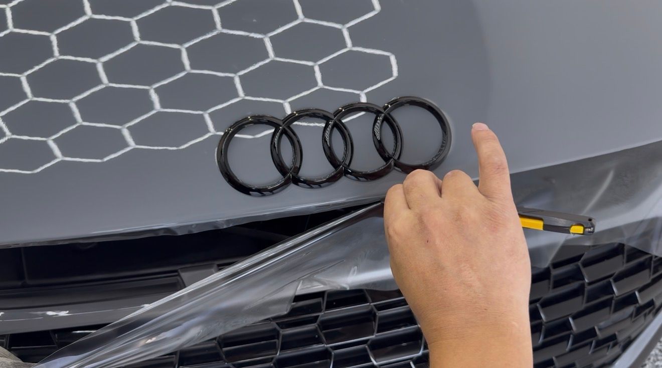 A hand uses a tool to carefully apply or trim a protective film around the black Audi logo on a gray car hood.