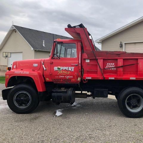 Red dump truck parked in a driveway, with red body and raised front bed, in a suburban neighborhood