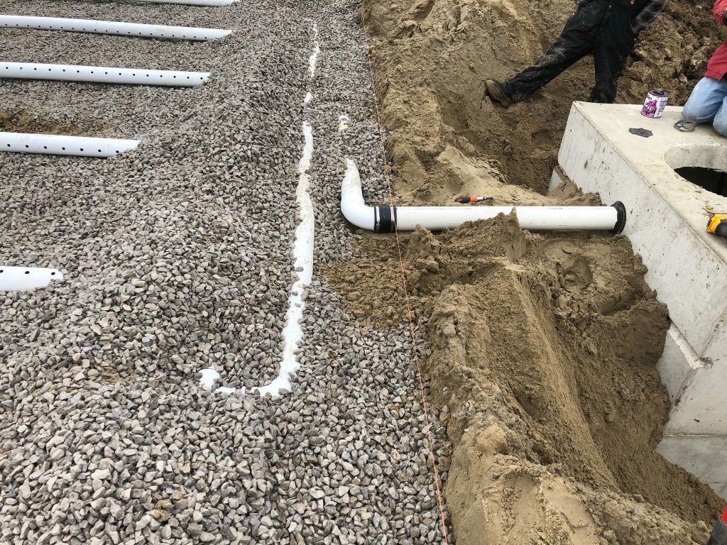 Gravel drainage trench with white PVC pipe and a small concrete footing beside a building.