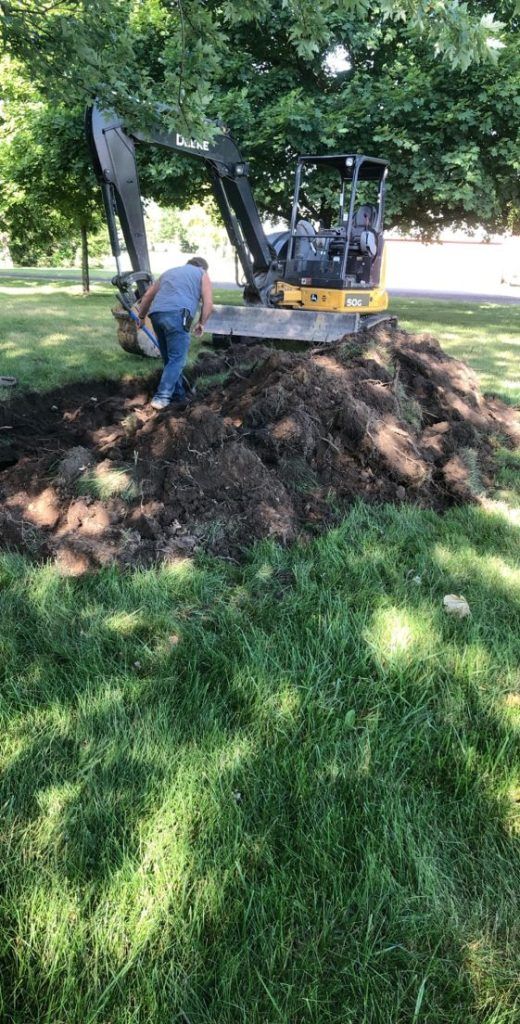 Man operating a mini excavator beside a large dirt pile in a grassy park under trees