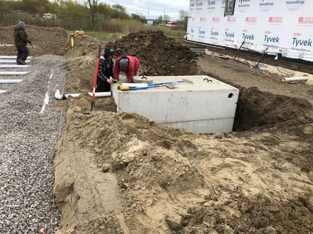 Construction worker beside a concrete utility box in a muddy excavation site with gravel and pipes.