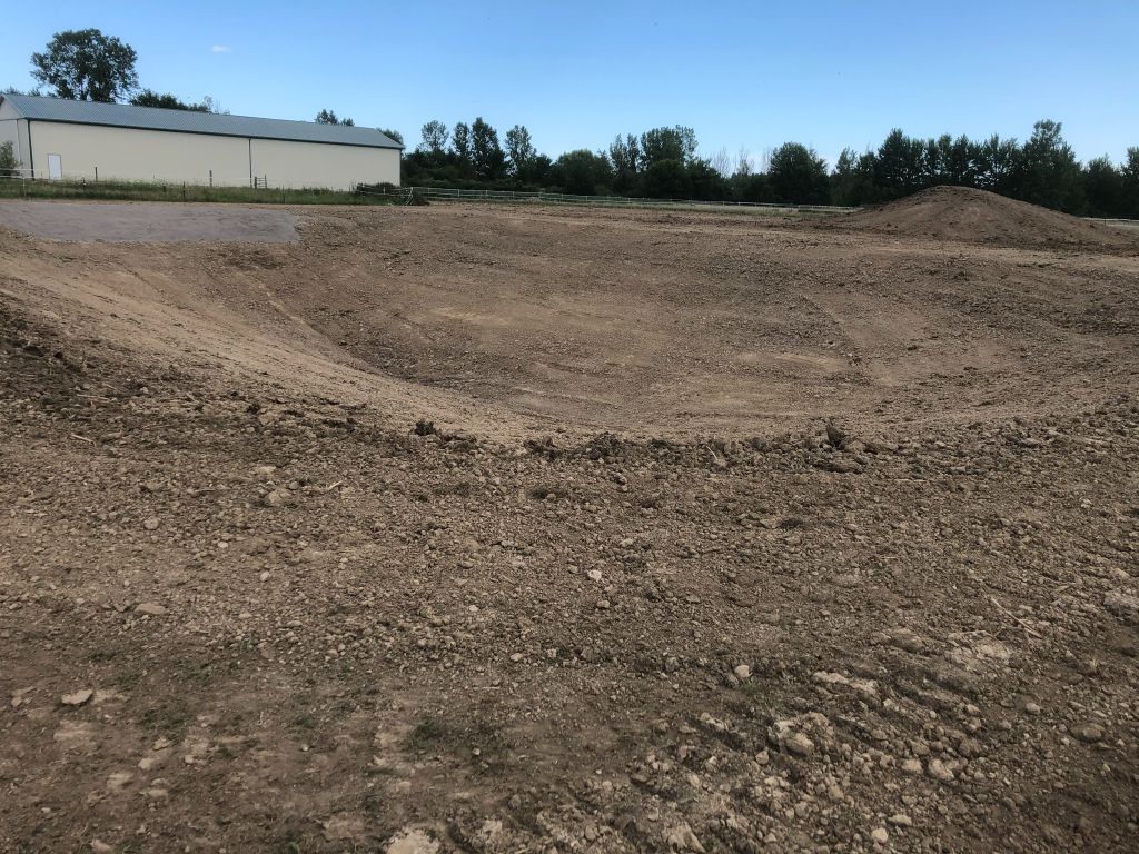 Bare dirt lot with a sloped embankment beside a white building and trees under a cloudy sky