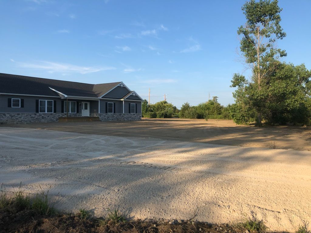 Single-story house with stone facade beside a large dirt lot and trees under a clear blue sky