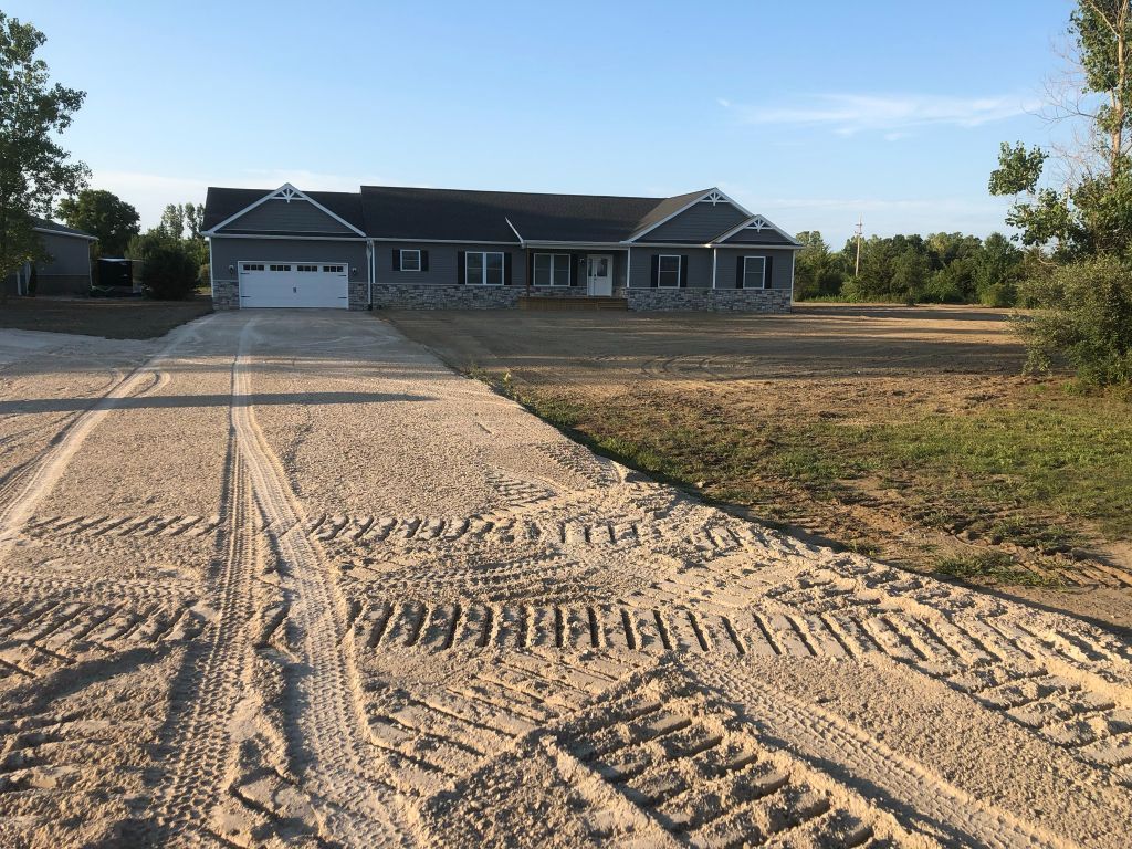 Single-story gray house with a long concrete driveway and freshly graded dirt yard under a clear sky