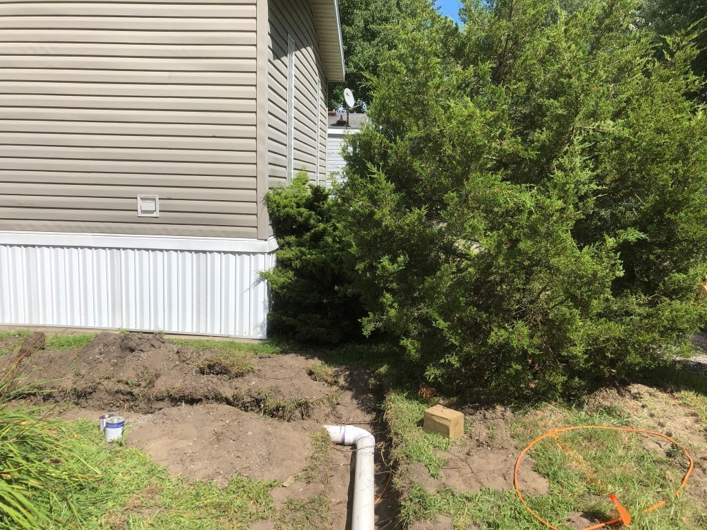 House side with beige siding, white skirting, a white downspout, and a green shrub beside a dirt slope.