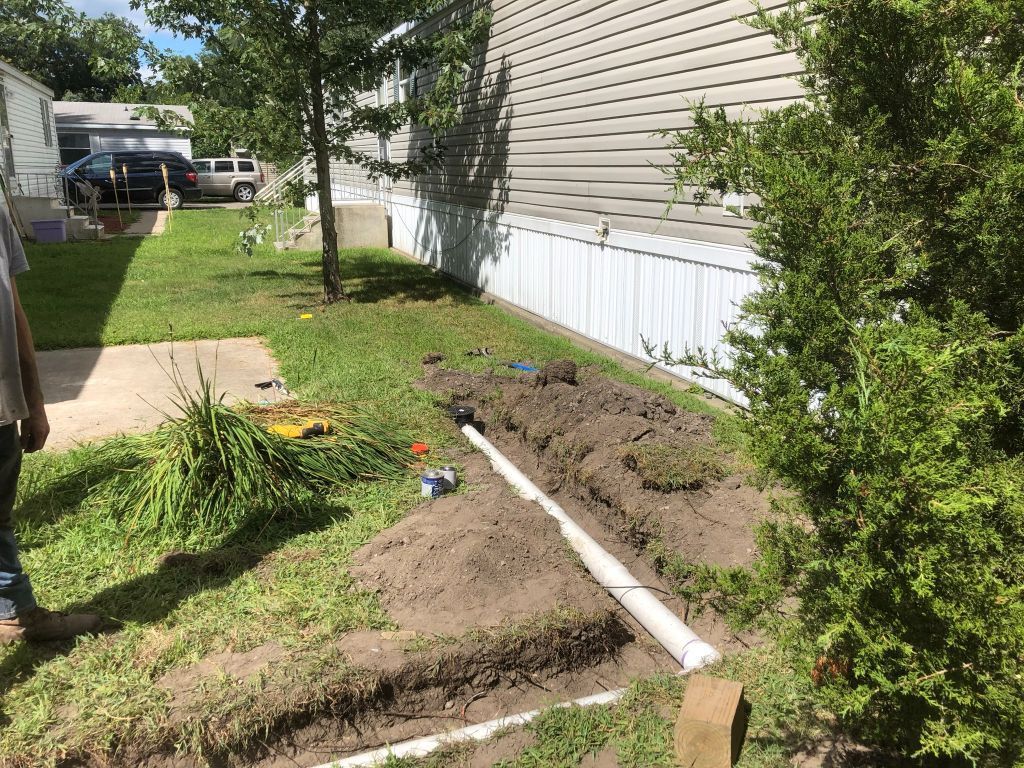 Backyard landscaping work with a trench, dirt piles, white pipe, and a house beside a grassy yard.