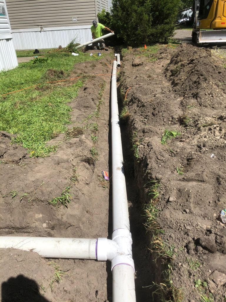 PVC drainage pipe trench beside a house, with dirt piles and a green lawn.
