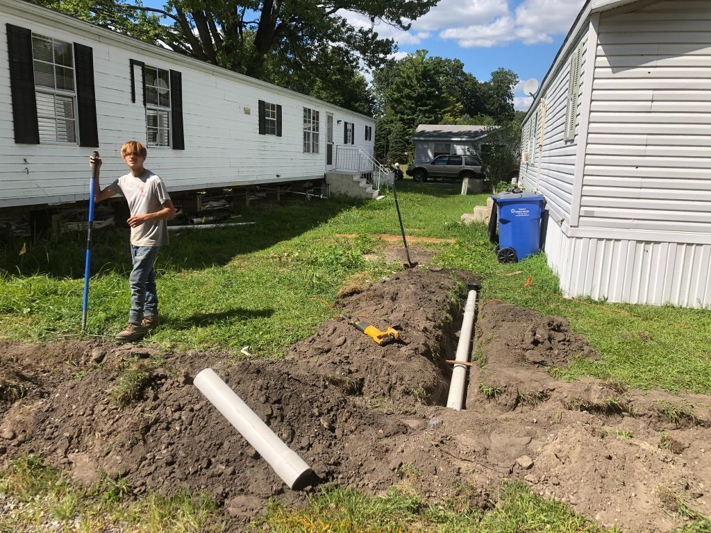 A person stands by a dug trench beside a white mobile home, with PVC pipes and dirt in the yard.