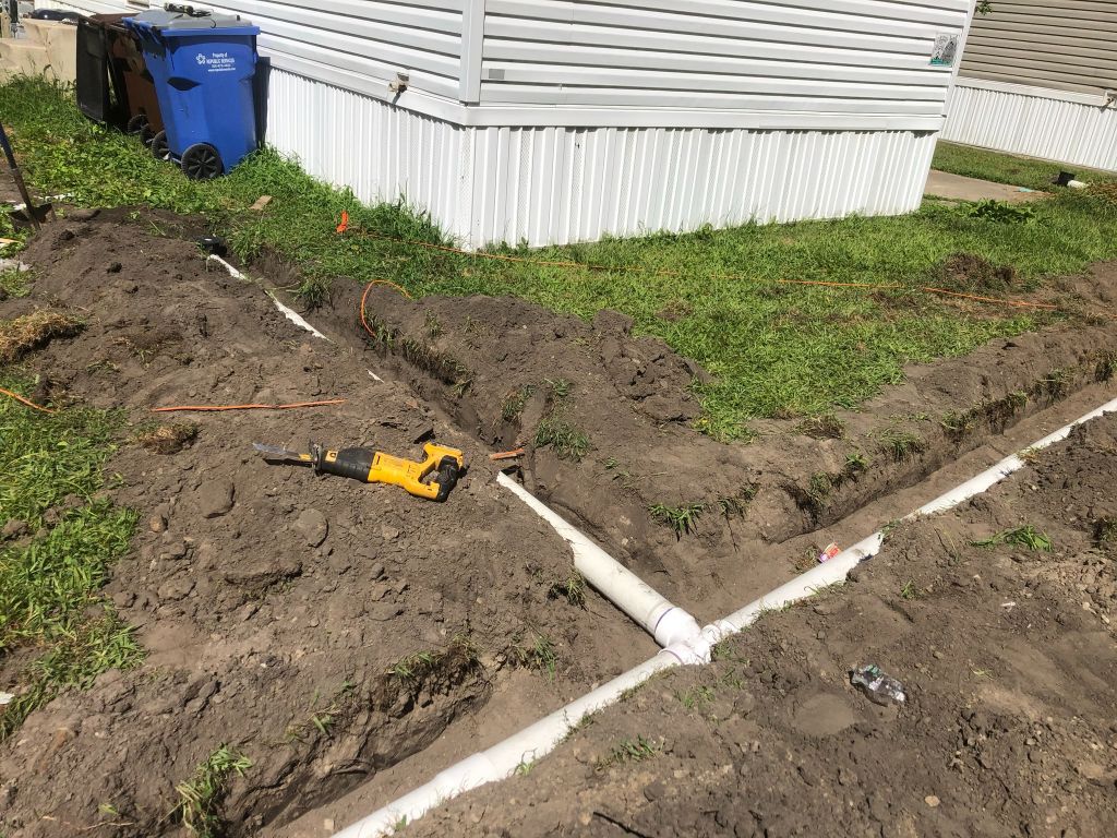 Excavation trench beside a mobile home with white pipes and a yellow utility tool on the ground