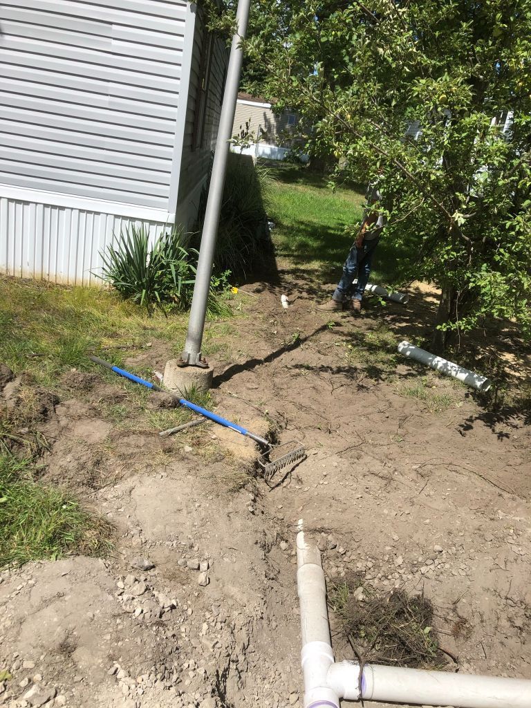 Backyard trench by a house with exposed white pipes and a blue shovel near dirt and grass.