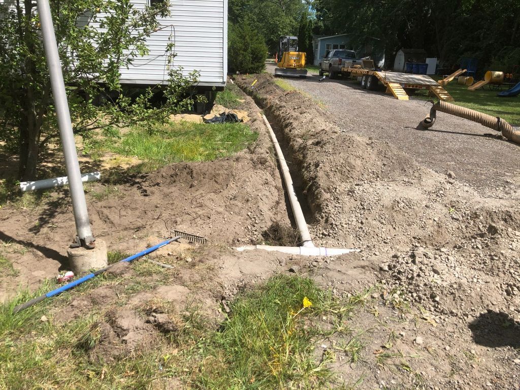 Construction site with gravel driveway, curb forms, and a white house beside a dirt yard