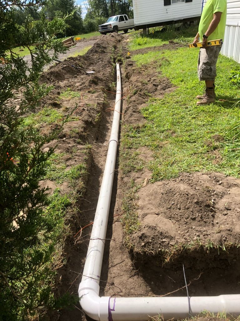 White PVC drainage pipe runs through a dug trench beside a house, with a person standing nearby.