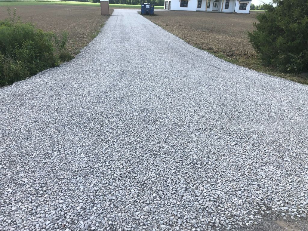 Fresh gravel driveway curving toward a house, bordered by grass and shrubs.