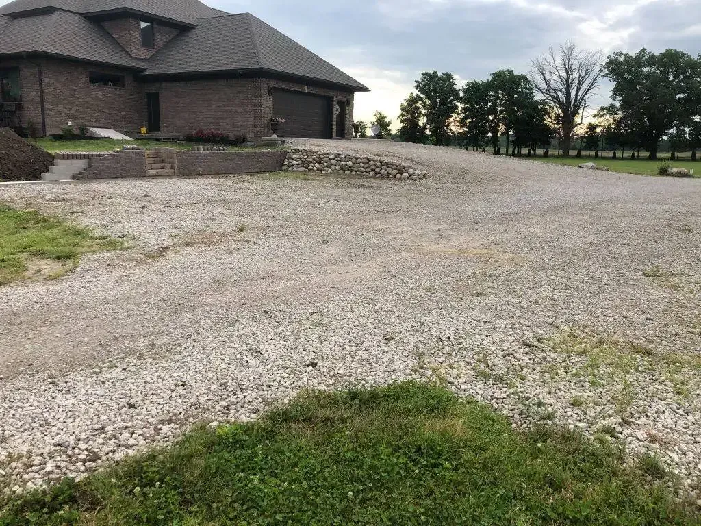 Gravel driveway beside a brick house with a grassy foreground and cloudy sky.
