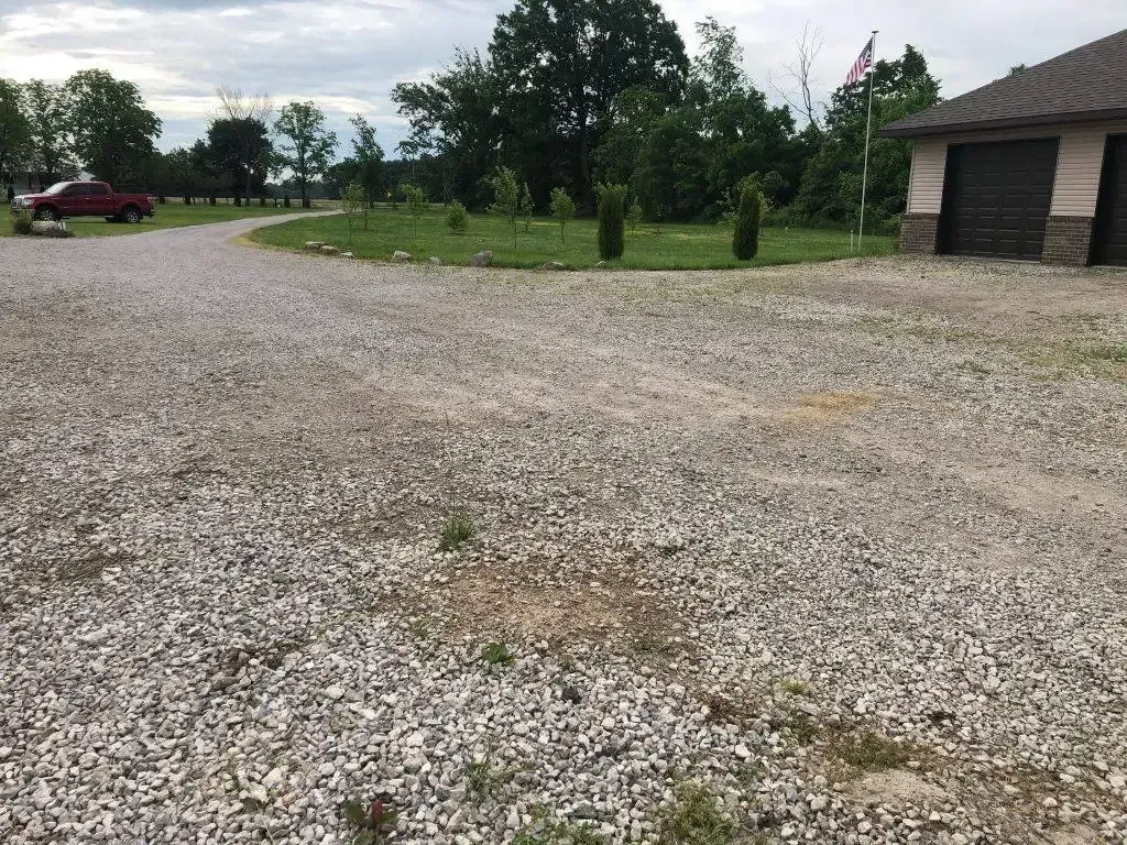 Gravel lot beside a small building, with trees, a parked red car, and cloudy sky.