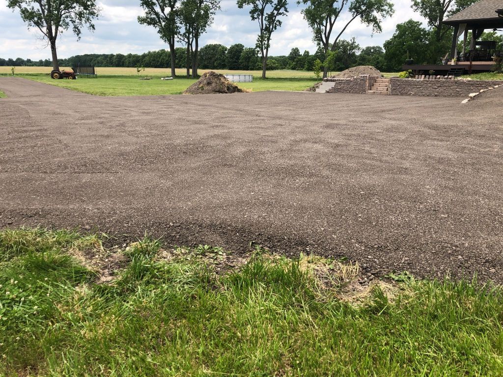 Gravel clearing in a park with grass, trees, and small piles of rocks under a cloudy sky