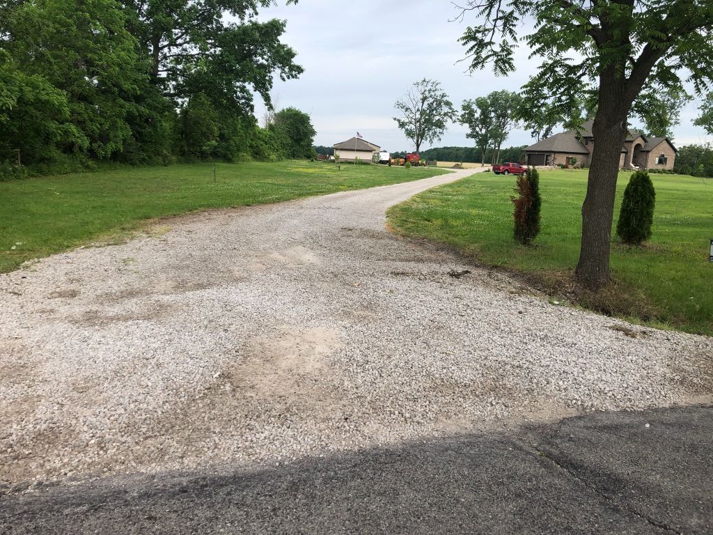 Gravel driveway curving through a grassy rural yard with trees and houses in the distance