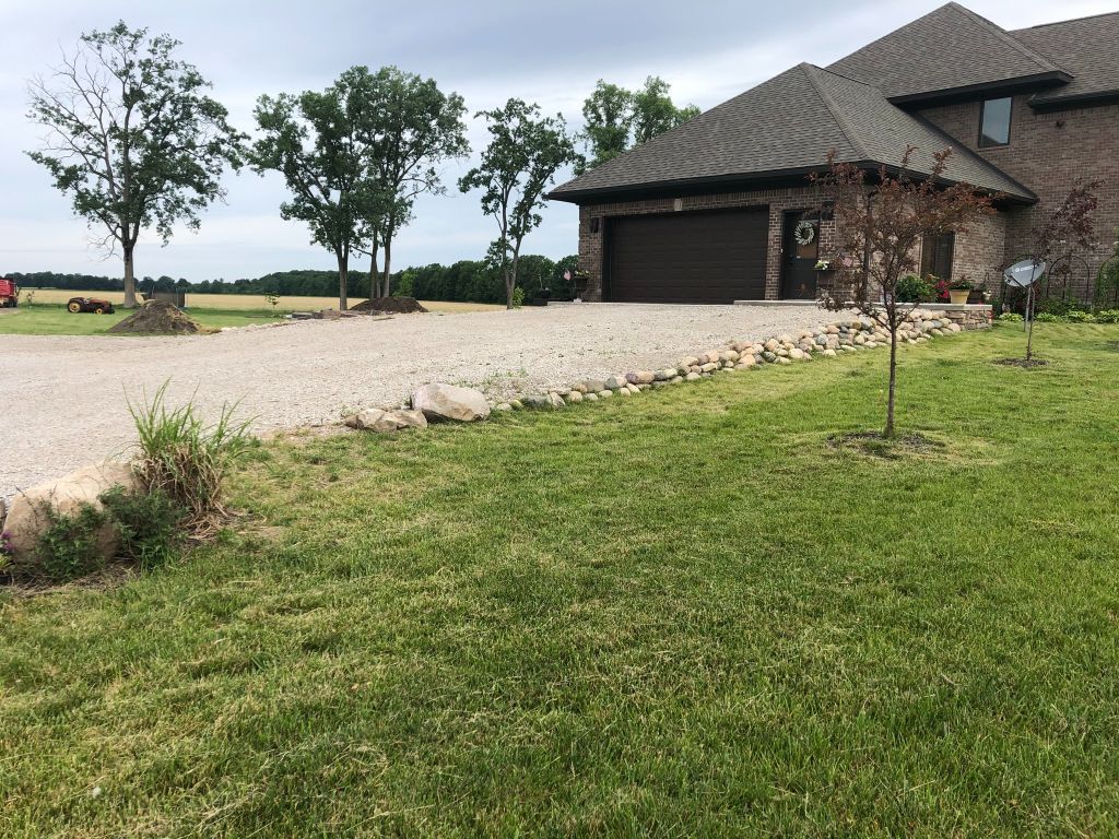 Gravel driveway beside a modern brick house with a dark roof, bordered by green lawn and young trees.